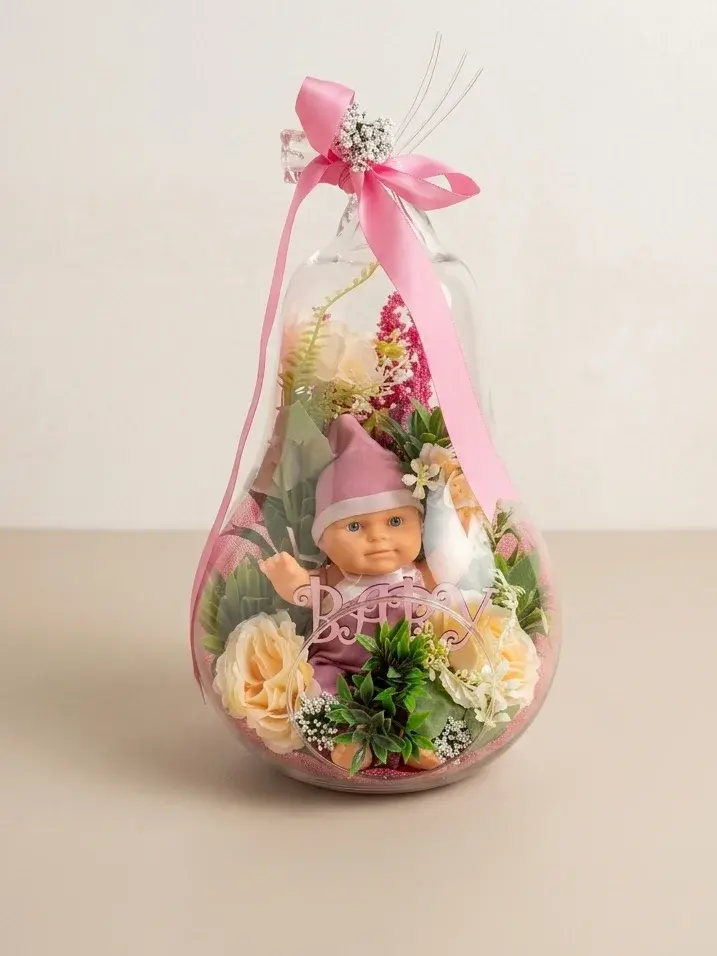 Armut biçimli cam fanus içinde pembe şapkalı bebek figürü, şampanya güller ve “Baby” yazısı; ağzında pembe saten fiyonk.
