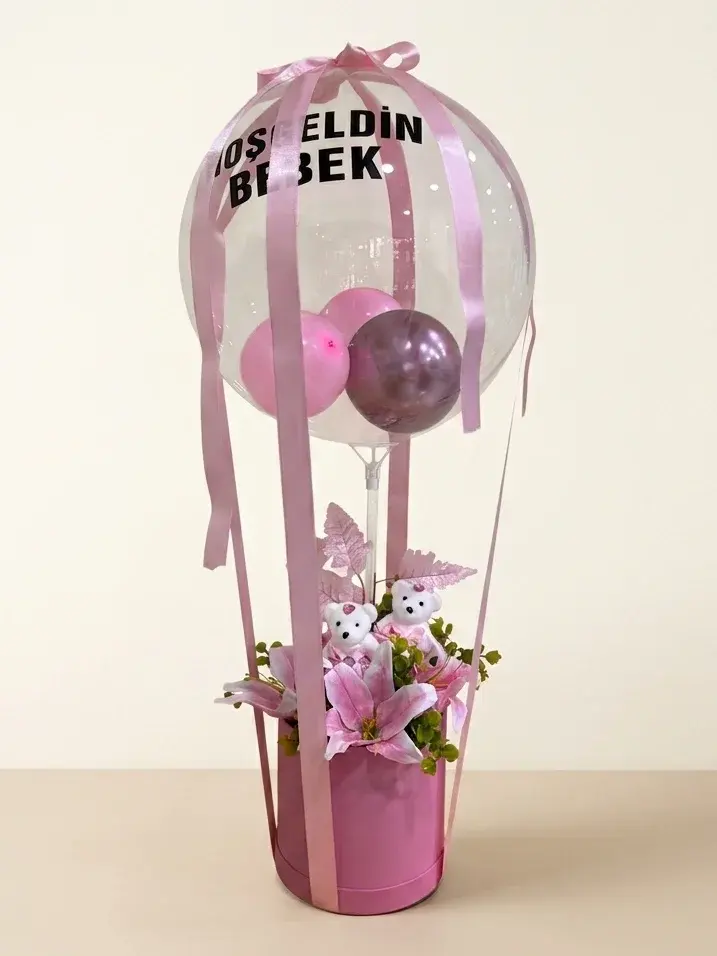 Pembe silindir kutu üzerinde “Hoşgeldin Bebek” yazılı şeffaf balon; içinde pembe balonlar, altında pembe lilyum ve peluş ayıcıklar.