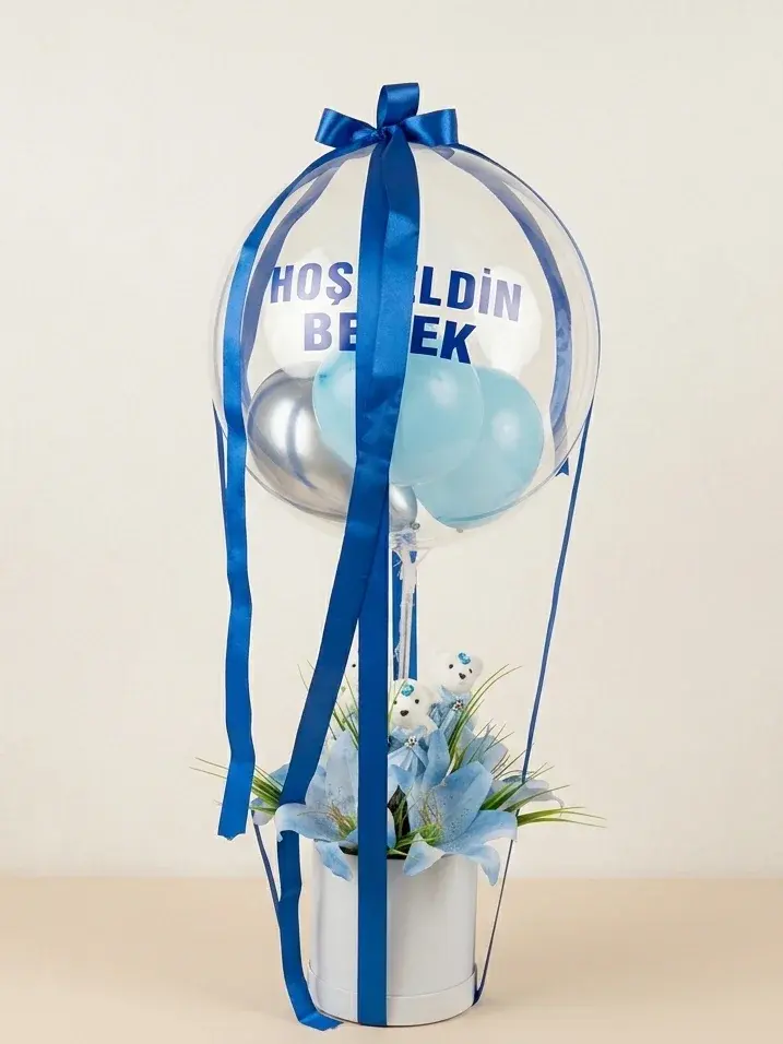 Beyaz silindir kutu üzerinde “Hoşgeldin Bebek” yazılı şeffaf balon; içinde mavi ve gümüş balonlar, lacivert kurdeleler, altında mavi lilyumlar.