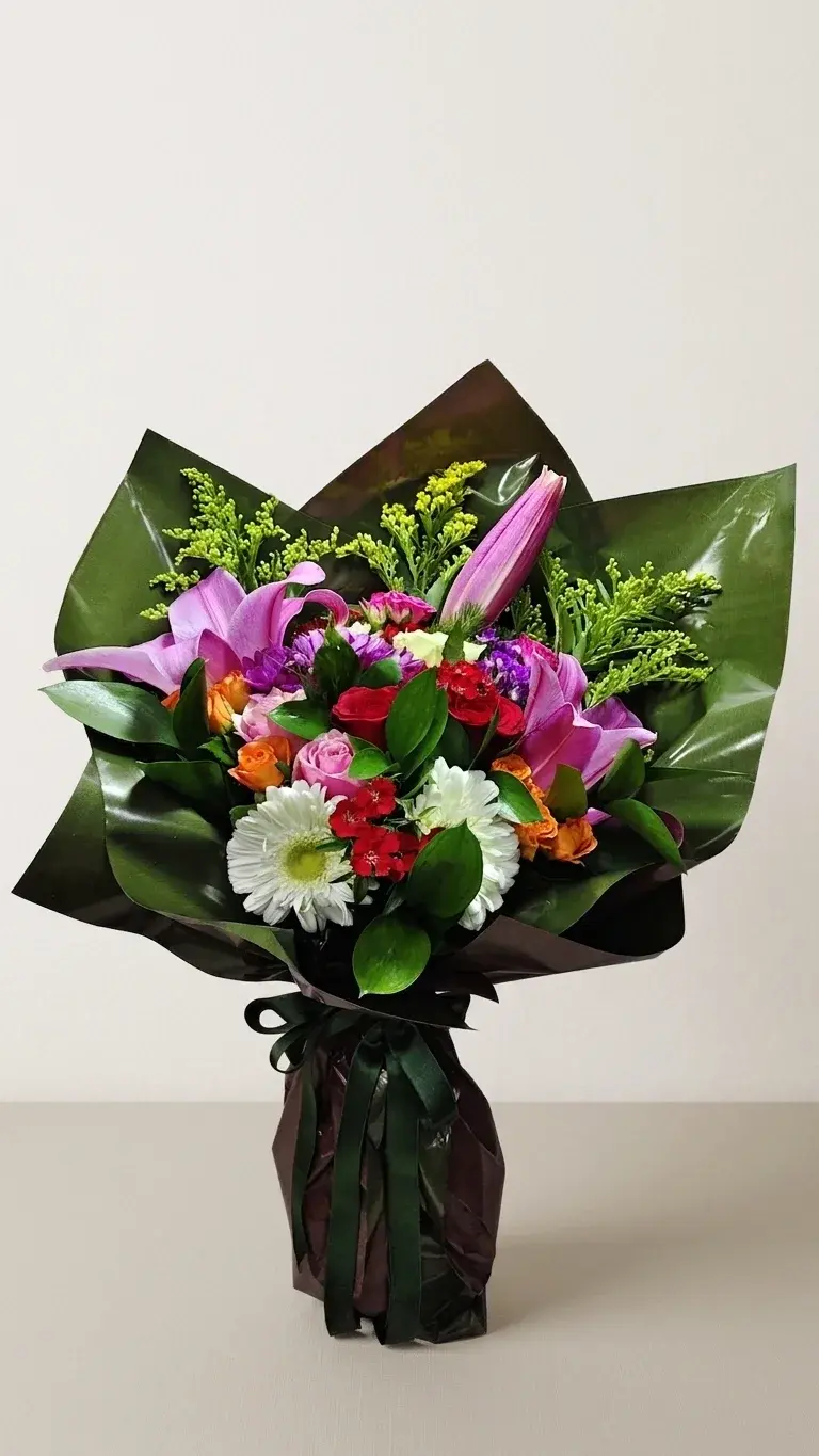 Koyu yeşil parlak ambalaj içinde lila lilyum, turuncu ve pembe güller, beyaz gerbera ve kırmızı karanfillerden karışık buket.